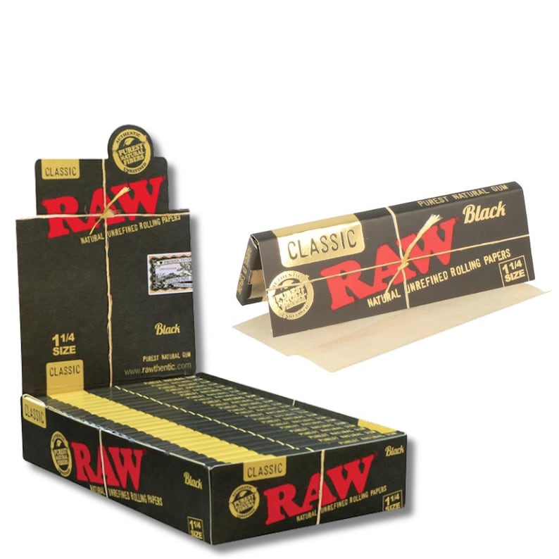 May include: A display box of black RAW Classic 1 1/4 size rolling papers. The box is black with red and gold accents, and the RAW logo is prominently displayed. A single pack of papers is shown next to the box.