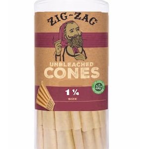 May include: A clear plastic cylinder containing unbleached cones. The packaging features the "Zig-Zag" logo with a bearded man smoking. The text "UNBLEACHED CONES" and "1 1/4 SIZE" are also visible. The cones are tan in color.