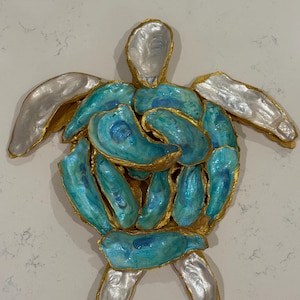 May include: A wall art sculpture shaped like a sea turtle, constructed from individual oyster shells. The central shell pieces are painted in vibrant shades of turquoise and blue, while the outer flippers and head are finished in silver.