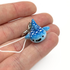 Peut inclure: Un petit porte-clés en forme de requin-baleine bleu avec des points blancs et un visage souriant. Le porte-clés a un fermoir argenté et une ficelle blanche. Le requin-baleine est bleu clair avec des nageoires et une queue bleu foncé.