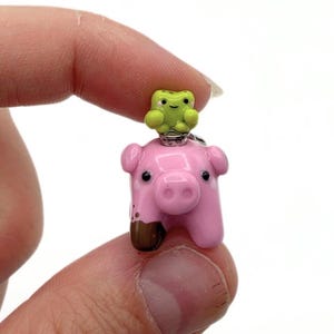 May include: A small, glossy charm featuring a pink pig with a brown patch on one leg and a tiny, light green frog perched on its head. The pig has a prominent snout, small ears, and black eyes, while the frog has a simple face with two black dots.
