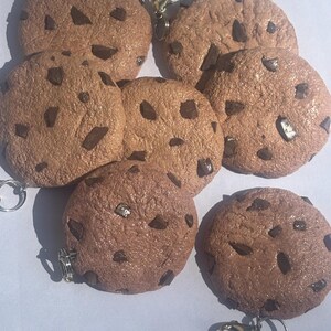 May include: Seven brown, chocolate chip cookie keyrings are displayed. Each keyring is a round, textured cookie with dark brown chocolate chips. The keyrings have a silver-coloured clasp for attaching to keys or bags.