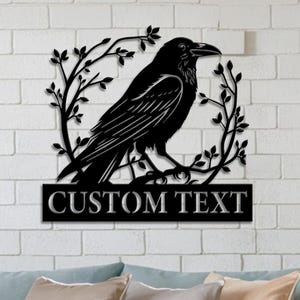 May include: Black metal raven wall art. The raven is perched on a branch within a circular wreath of leaves. The words "CUSTOM TEXT" are displayed on a rectangular plaque below the raven. The art is mounted on a white brick wall.