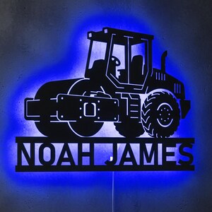 Peut inclure: Silhouette noire d'un rouleau compresseur routier avec rétroéclairage LED bleu. Le nom "NOAH JAMES" est affiché en dessous. L'image est fixée sur un mur gris.
