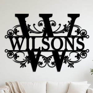 Puede incluir: Arte mural de metal negro con el nombre "Wilsons" entre dos letras "W" grandes. Las letras y el nombre están conectados por adornos florales decorativos. La obra de arte está montada en una pared blanca sobre un sofá blanco.