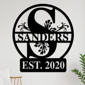 Puede incluir: Arte mural de metal negro con el nombre "Sanders" dentro de un marco circular, con una gran letra "S" y detalles florales. El diseño también incluye "EST. 2020". La obra de arte está montada en una pared blanca.