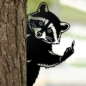 May include: Black metal raccoon silhouette peeking from behind a tree trunk, giving the middle finger. The raccoon has white markings around the eyes and on the face. The tree trunk is brown and textured.