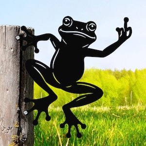 May include: Black metal frog silhouette climbing a wooden post, giving the middle finger. The frog has large eyes and is set against a backdrop of green grass and a blue sky. Decorative garden art.