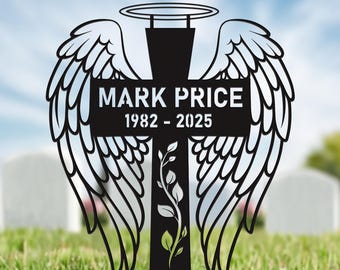 May include: Black metal memorial cross with angel wings, a halo, and the name "MARK PRICE" with the years "1982 - 2025". The cross features a floral design and is set in a grassy cemetery.