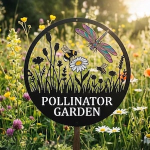 Puede incluir: Un letrero de jardín circular de metal negro con un diseño recortado de una libélula, abejas y flores. El texto "POLLINATOR GARDEN" está centrado en letras blancas en la parte inferior. El letrero está montado en una estaca de madera.