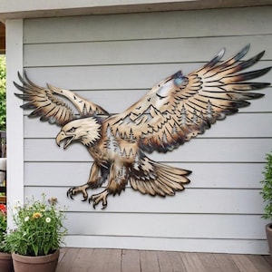 Op de afbeelding: Een groot metalen wandkunstobject in de vorm van een vliegende Amerikaanse zeearend met wijd gespreide vleugels. De adelaar heeft een uitgesneden ontwerp dat een bergketen en een dennenbos binnen de vleugels en het lichaam weergeeft.
