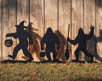 May include: Black metal Halloween yard art depicting three Bigfoot silhouettes. One wears a witch's hat and carries a jack-o'-lantern bucket. Another holds a cauldron, and the third waves while holding a broom. The figures stand on grass in front of a wooden fence.