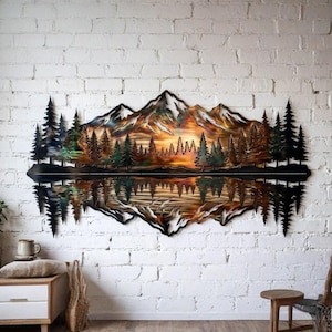 May include: Metal wall art showing a mountain scene with a lake reflection. The artwork features a sunset with orange and yellow hues, silhouetted trees, and snow-capped mountains. The piece is mounted on a white brick wall.