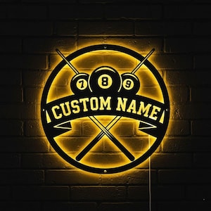 Può includere: Un'insegna rotonda, nera e dorata, illuminata a tema biliardo. L'insegna presenta tre palle da biliardo, stecche incrociate e la scritta "CUSTOM NAME" su uno striscione. I numeri 7, 8 e 9 sono visibili sulle palle da biliardo.