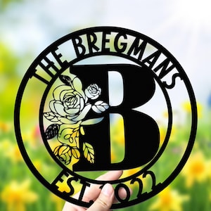 Puede incluir: Letrero de nombre familiar de metal negro con el texto "THE BREGMANS" y "EST 2022" en un diseño circular. Una gran letra "B" está en el centro, con un diseño floral. El letrero se sostiene frente a un fondo borroso amarillo y verde.