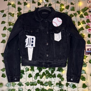 May include: Black denim jacket with a baseball patch and a white patch on the chest pocket. The jacket features a white patch with a stylized letter "D" on the left chest. The jacket is buttoned up and hanging on a black hanger.