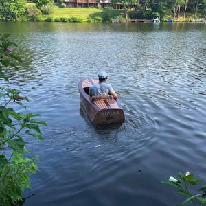 以下が含まれることがあります： 湖に浮かぶ小さな木製のボートに乗った人。ボートは茶色で、明るい色の木の甲板があり、側面に「STELLA」という名前が見えます。その人は帽子と明るい色のシャツを着ています。背景には木と建物が写っています。