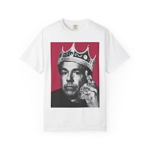 May include: White t-shirt featuring a black and white portrait of a man wearing a crown and multiple rings, set against a red background. The image is centered on the shirt, showcasing the graphic design. The shirt has a classic crew neck.