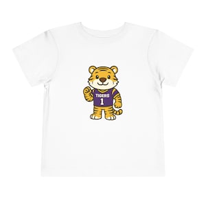 May include: White toddler t-shirt featuring a cartoon tiger wearing a purple and gold jersey with the word "Tigers" and the number "1". The tiger is smiling and giving a thumbs up.