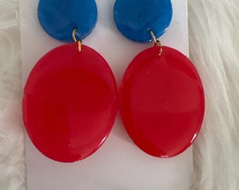 May include: A pair of vibrant earrings featuring a blue circular top and a red oval bottom. The earrings are connected by a small gold-colored loop. The earrings are displayed on a white card.