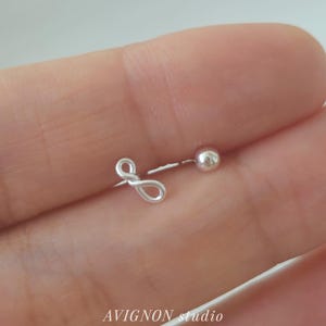 Op de afbeelding: Een zilveren oorbel met een kleine bal en een lusvormig ontwerp. De oorbel wordt in een hand gehouden, met de tekst "AVIGNON studio" zichtbaar in de afbeelding.