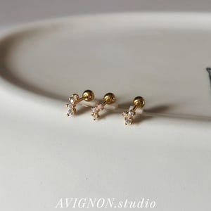 May include: Three gold-tone stud earrings, each featuring a curved bar design set with three small clear stones and a round ball backing. The earrings are arranged in a row, showcasing a metallic finish and delicate, sparkling stone accents.