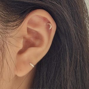 以下が含まれることがあります： 2つのシルバーのイヤリングが付いた耳のクローズアップ。1つは小さなフープイヤリング、もう1つは三日月型です。「AVINGNON studio」の文字が画像の下部に表示されています。