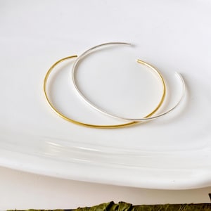 May include: Two open-ended bangles, one gold and one silver, sit on a white, oval plate. The bangles are thin and circular, with a polished finish. The text "AVIGNON studio" is printed in a light grey colour at the top of the image.