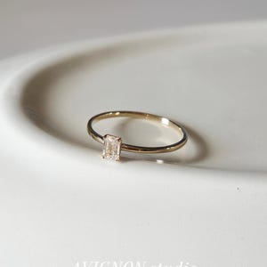 May include: A delicate gold ring with a rectangular, clear gemstone. The ring is set on a white surface and is the focal point of the image. The text "AVIGNON.studio" is at the bottom.