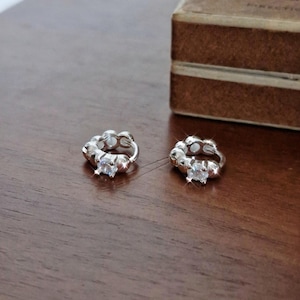 Könnte beinhalten: Zwei silberfarbene, offene Creolen mit einem Cluster-Design und einem quadratischen, klaren Edelstein in der Mitte. Die Ohrringe liegen auf einer dunklen Holzoberfläche, im Hintergrund befindet sich eine braune Schachtel.