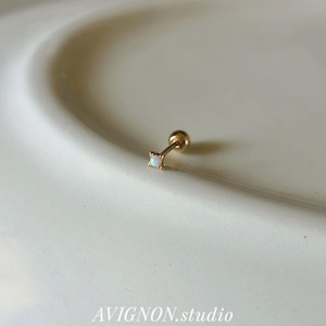 May include: A gold-toned stud earring featuring a small, round, iridescent white opal stone set in a four-prong star-shaped mount. The earring has a thin post and a spherical back closure. The text AVIGNON.studio is visible at the bottom.
