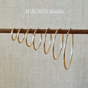 Puede incluir: Una colección de pendientes de aro dorados de varios tamaños se muestra en una varilla de madera. Los pendientes están dispuestos de menor a mayor, mostrando su diseño circular y acabado brillante. El texto "AVIGNON studio" es visible en el fondo.