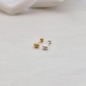 May include: Two small stud earrings, one gold and one silver. Each earring features an oblong shape at one end and a small ball at the other, connected by a straight post. The earrings are presented on a white surface. The earrings are approximately 1 cm in length.