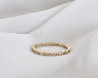 Può includere: Un delicato anello d'oro con una fila continua di piccole pietre preziose trasparenti. L'anello è posto su uno sfondo di tessuto bianco strutturato, creando un'estetica semplice ed elegante. Il design dell'anello è minimalista.