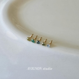 Könnte beinhalten: Sechs kleine Ohrstecker mit farbigen Edelsteinen in Grün- und Blautönen sind auf einer weißen Oberfläche angeordnet. Die Ohrringe haben goldene Stifte. Der Text "AVIGNON studio" befindet sich unten.