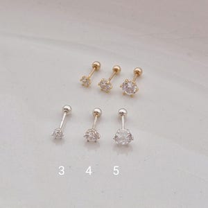 以下が含まれることがあります： 透明なキュービックジルコニアの石が付いたスタッドピアス6個。3つはゴールドトーン、3つはシルバートーンです。ピアスには3、4、5の番号が付けられています。ピアスは明るい色の表面に表示されています。