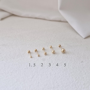 Könnte beinhalten: Fünf goldene Kugelohrstecker in verschiedenen Größen sind in einer Reihe angeordnet. Die Größen sind mit 1,5, 2, 3, 4 und 5 beschriftet. Die Ohrringe werden auf einer weißen Oberfläche mit einem weichen, neutralen Hintergrund präsentiert.
