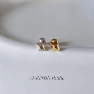 以下が含まれることがあります： 幾何学的なピラミッドデザインのスタッドピアス2つ。1つはシルバー、もう1つはゴールドです。白い表面に表示されています。「AVIGNON studio」の文字が下に印刷されています。