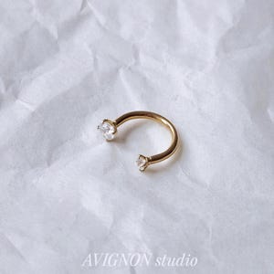 Op de afbeelding: Een goudkleurige ring met twee heldere, ronde edelstenen. De gebogen ring ligt op een witte, getextureerde ondergrond. De tekst "AVIGNON studio" is in donkergrijs gedrukt.