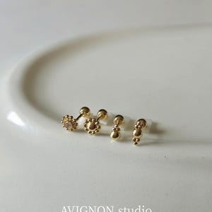May include: Four gold-toned stud earrings with flat-back posts. Two feature a flower-like design with small spheres, one with a central clear stone. The other two have vertical arrangements of small spheres. The text AVIGNON.studio is visible.