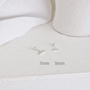 Puede incluir: Dos pequeños pendientes de botón plateados con un diseño de barra. Un pendiente mide 7 mm de largo y el otro 5 mm. Los pendientes se muestran sobre una superficie blanca con las medidas impresas debajo.