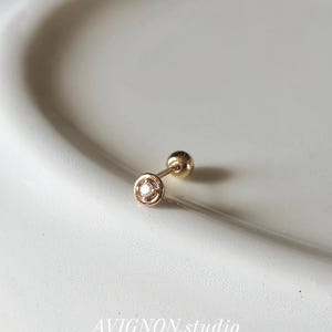 May include: A gold stud earring featuring a small, circular setting with a central clear gemstone. The earring has a thin post and a spherical gold backing. The text AVIGNON.studio is visible at the bottom of the frame in white lettering.