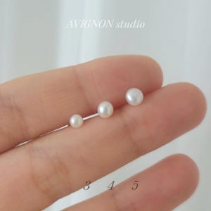 May include: Three small, white pearl stud earrings of varying sizes are displayed. The earrings are arranged in a row, with the smallest on the left and the largest on the right. The text "AVIGNON studio" is visible.
