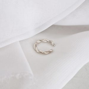 Puede incluir: Un pendiente de oreja plateado con un diseño de cuerda trenzada. La joya se encuentra sobre un fondo de tela blanca, creando una estética minimalista. El pendiente de oreja es un accesorio moderno.