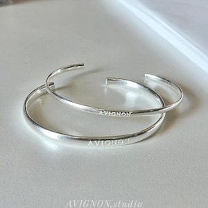 May include: Two silver bangles with an open design. The word "AVIGNON" is engraved on the outer surface of each bangle. The bangles are displayed on a light-coloured surface. The text "AVIGNON.studio" is visible below the bangles.