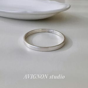 May include: A silver-colored bangle bracelet with a flat design, resting on a light-colored surface. The bracelet is circular and appears to be made of metal. A white, oval-shaped dish is in the background. The text "AVIGNON studio" is at the bottom.