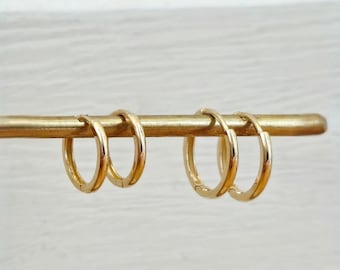 May include: Two pairs of small, polished gold hoop earrings hanging on a thin, horizontal gold-toned rod. The earrings feature a simple, circular design with a smooth, reflective surface. The text AVIGNON studio is printed in white at the bottom.
