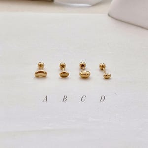 May include: Four gold-colored stud earrings with rounded, organic shapes. Each earring has a small, round post. The earrings are labeled A, B, C, and D. The earrings are displayed on a white surface.