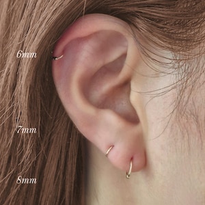 Puede incluir: Primer plano de una oreja con tres pequeños pendientes de aro dorados. Las medidas son 6 mm, 7 mm y 8 mm. El texto "AVIGNON studio" está en la parte inferior. El pelo castaño enmarca la oreja.