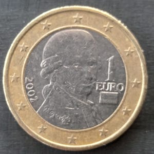 Puede incluir: Una moneda de un euro de 2002. La moneda presenta un retrato de un hombre con peluca empolvada y el número "1 EURO". La moneda tiene un anillo exterior dorado con estrellas.